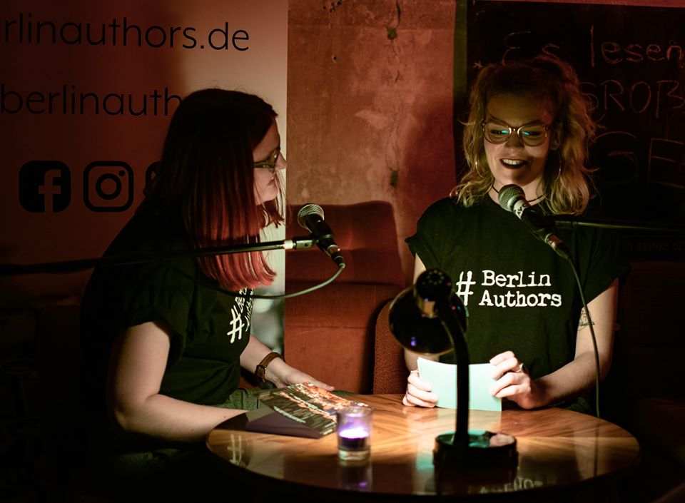 Das Bild zeigt Katha, die Sophie bei einer Lesung interviewt. Die beiden sitzen an einem Tisch bei dämmriger Beleuchtung. Sie tragen schwarze T-Shirts, auf denen in weiß "#BerlinAuthors" steht.