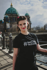 Frau mit braunen Haaren, rotem Bandana und schwarzem T-Shirt steht vor dem Berliner Dom.